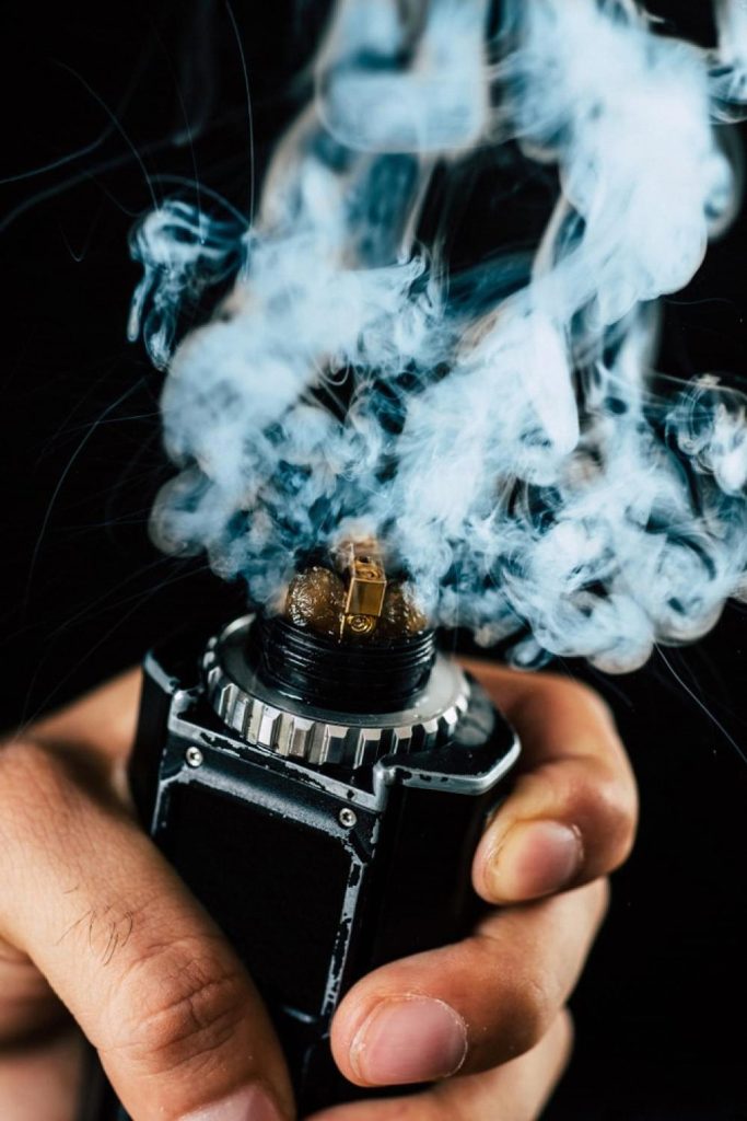 Hand holding an advanced vape mod with a visible coil producing a large cloud of vapour