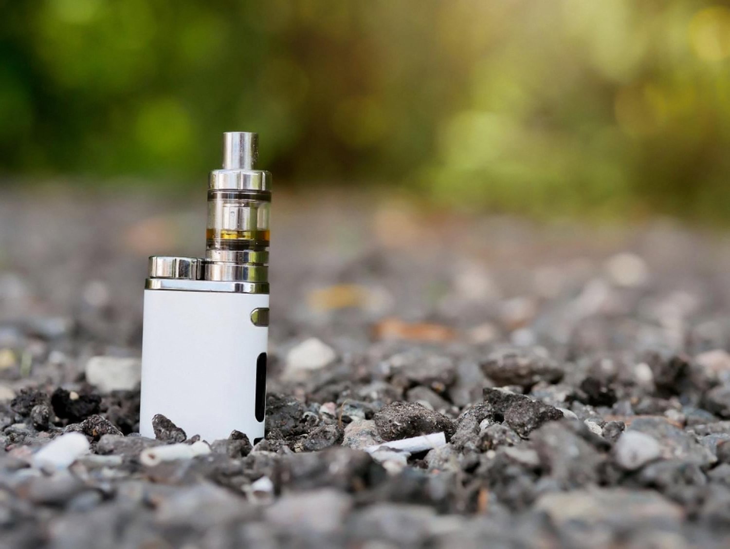 Compact white pod-style vape device standing on gravel with greenery in the background